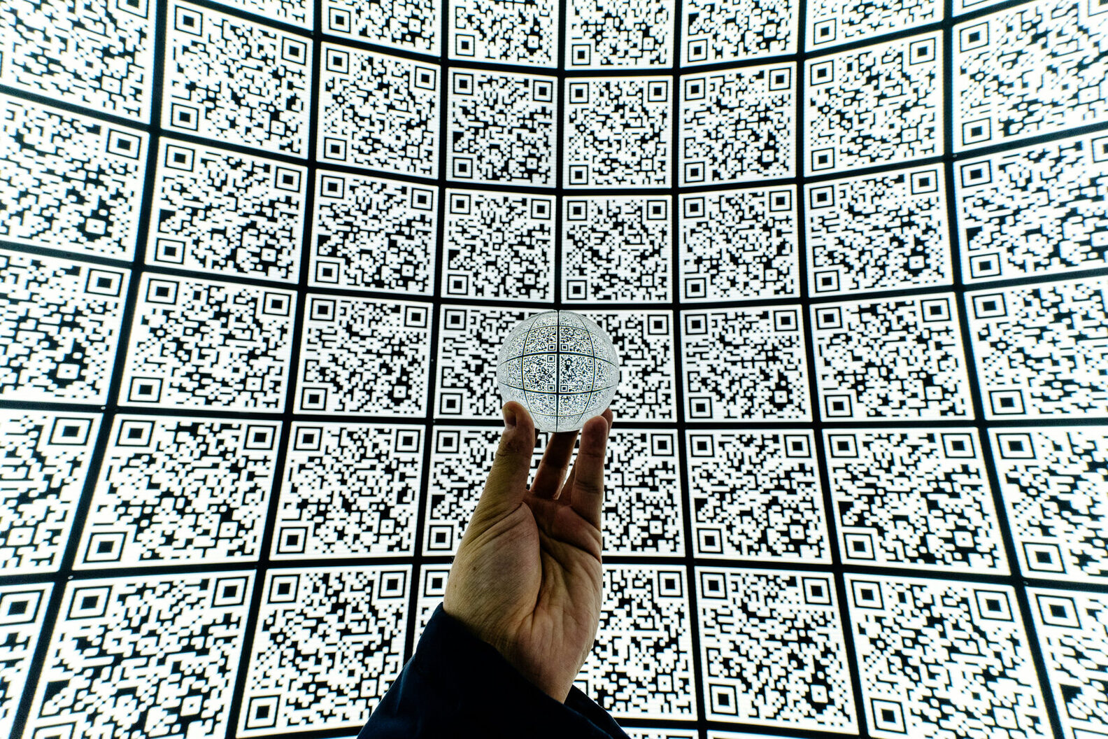 Una mano regge una sfera davanti a una grande superficie coperta di codici QR
